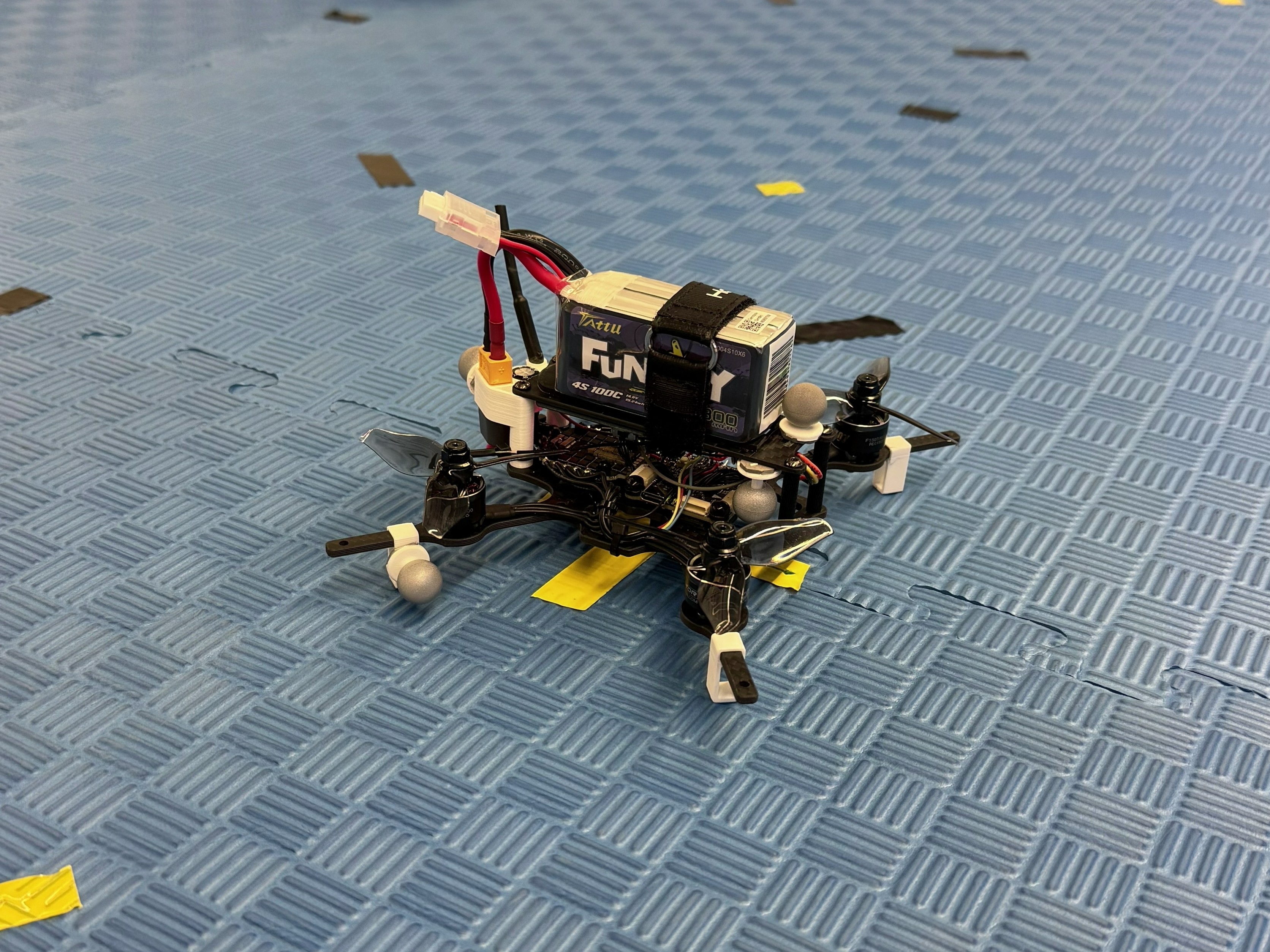 A small custom quadrotor on the arena floor, with motion-capture markers